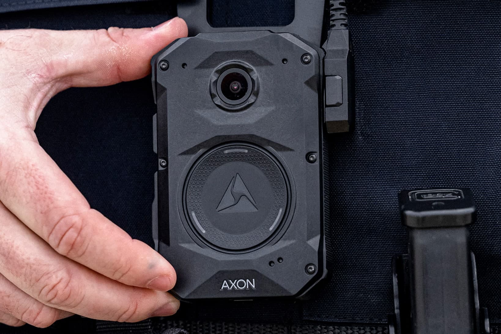 An Axon Body 4 camera on an officer's vest, a hand holding it by its edge