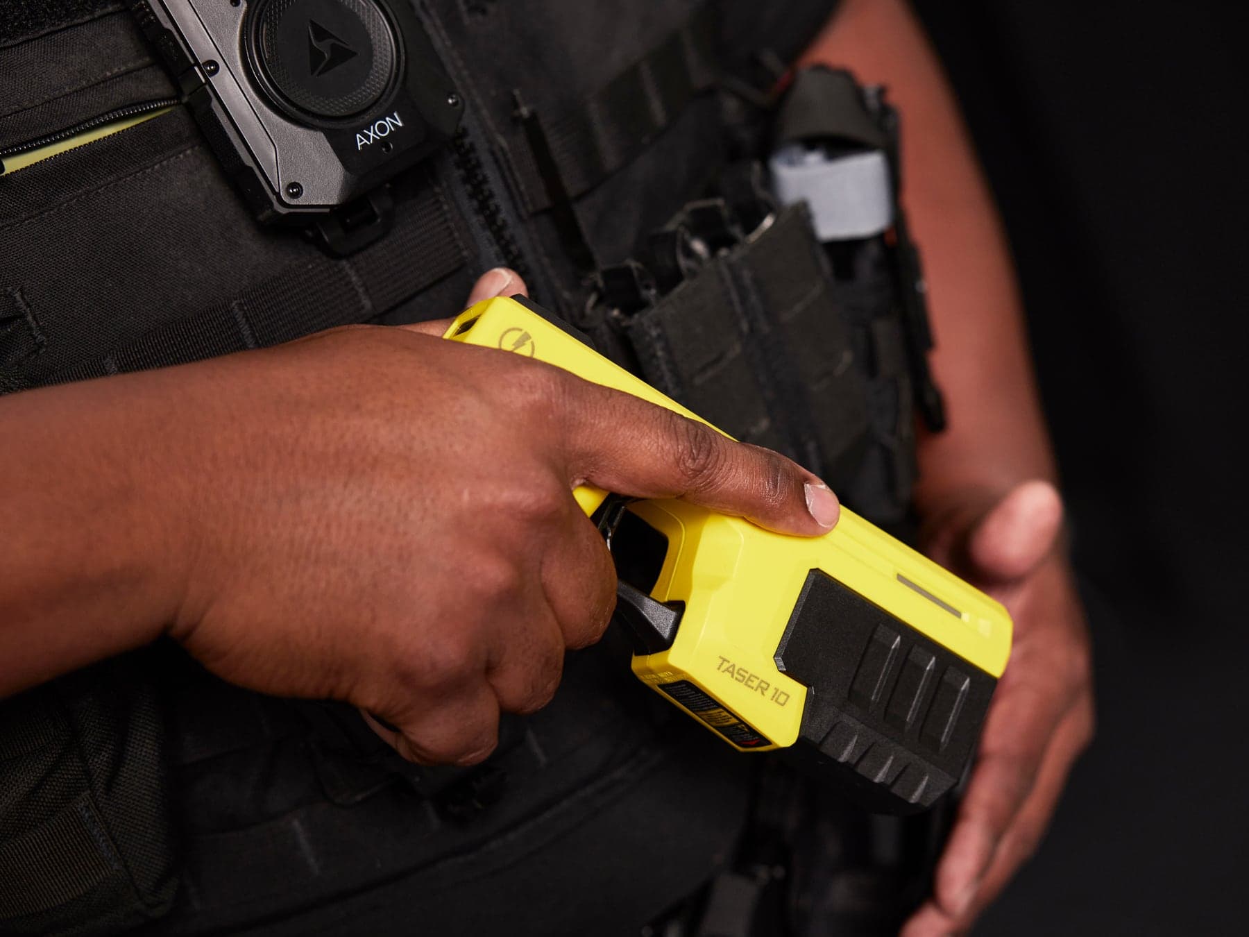 A TASER 10 energy weapon held in front of a vest, with an Axon body-worn camera above