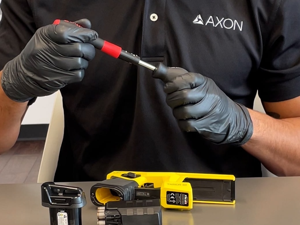 A gloved technician in an Axon shirt services a TASER 10 energy weapon on a bench, its parts set out beside it.