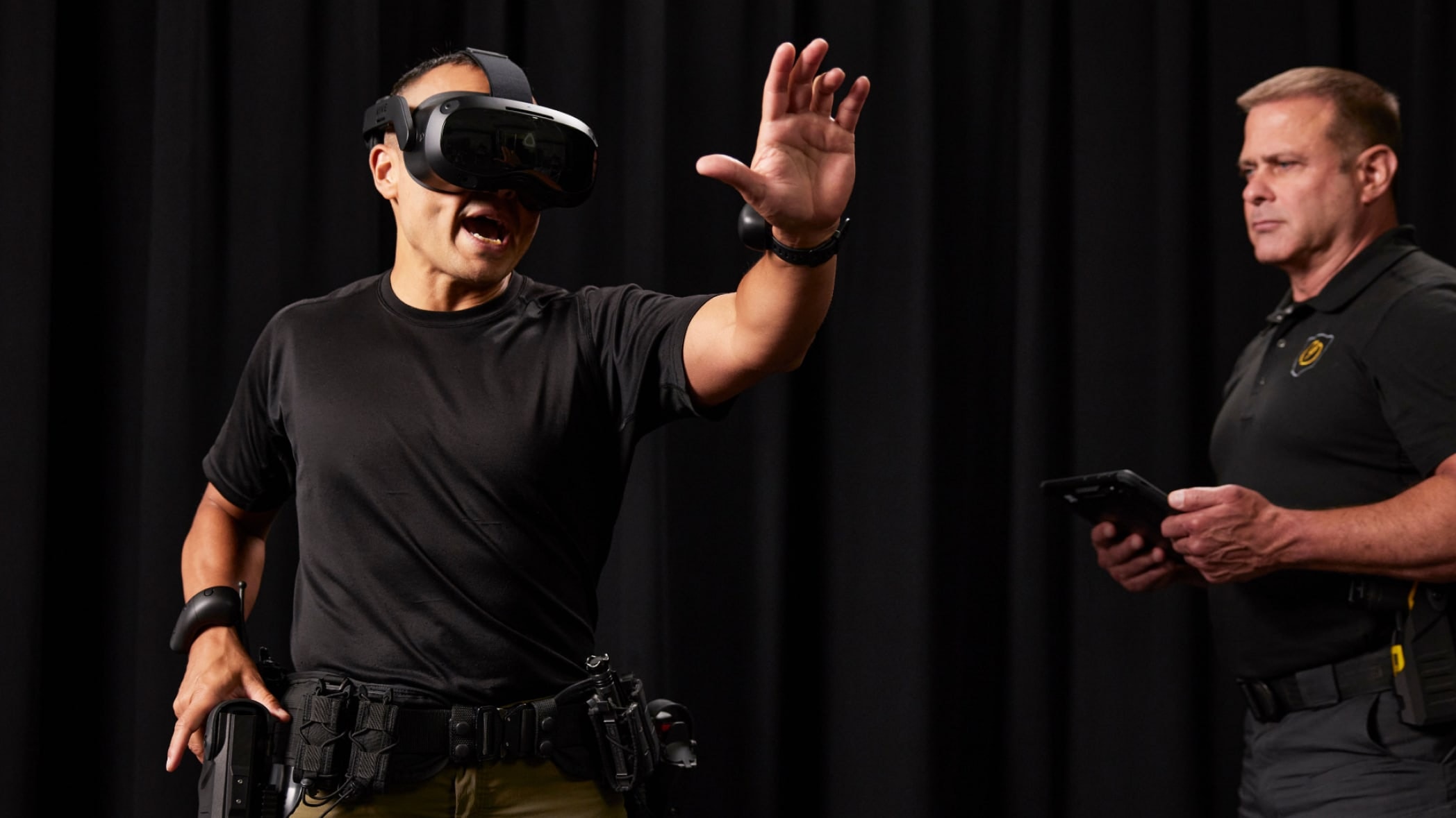 Training with the Axon VR training system: a trainee in a VR headset raises an open hand and gives a verbal warning while an instructor follows on a tablet.